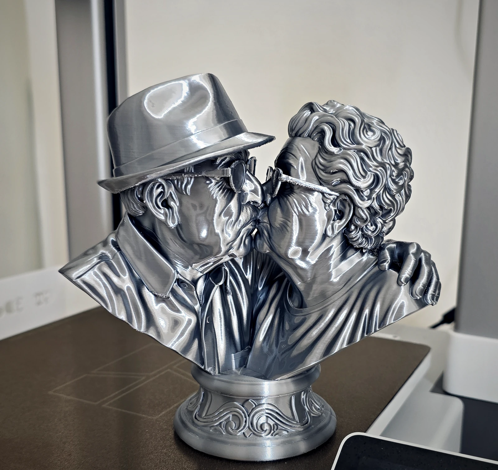Escultura em prata de um casal de idosos se beijando, ele de chapéu e óculos, ela de cabelos cacheados