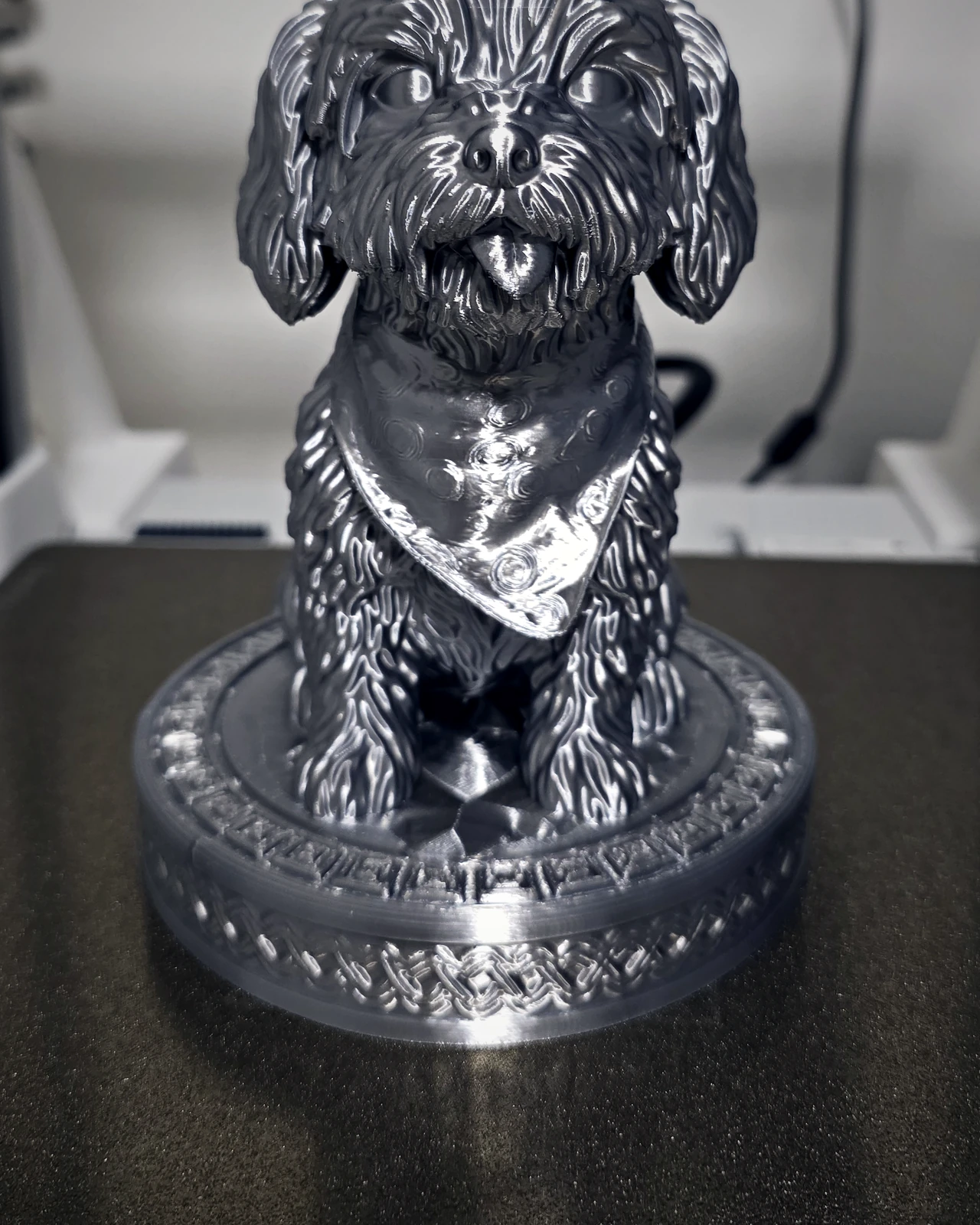Escultura prateada de um cachorrinho peludo de lenço no pescoço, sobre base decorada
